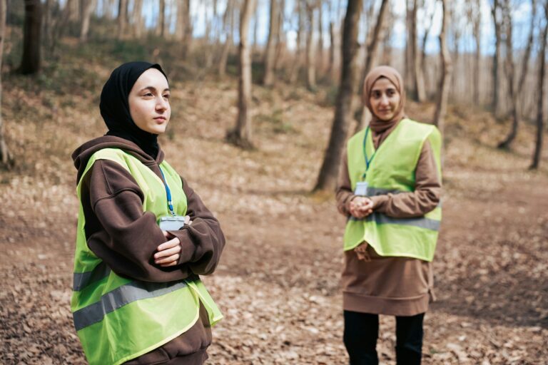 Two women wearing head coverings and bright safety vests standing in a forest, representing community care, environmental stewardship, and resilience